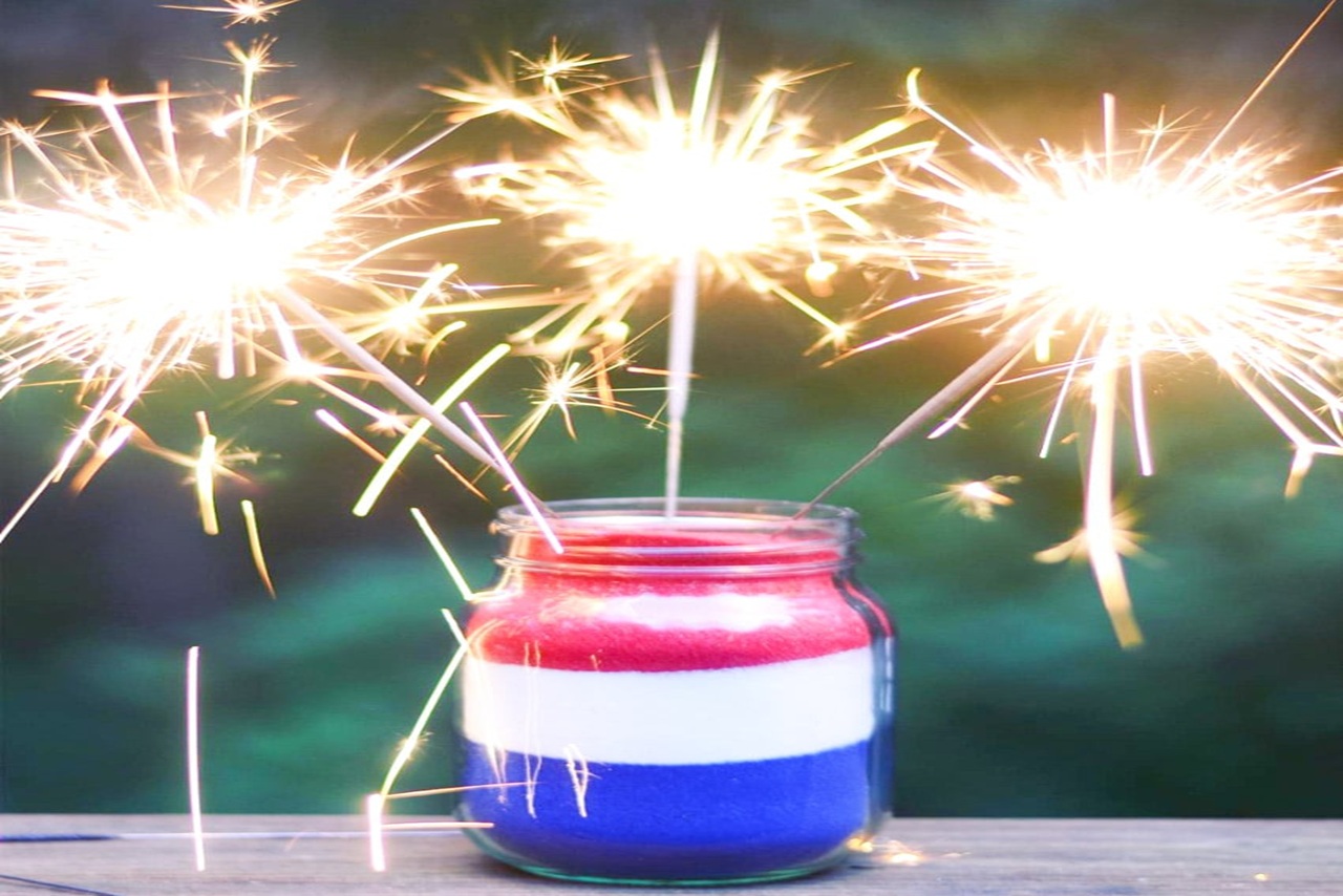 red, white and blue sand layered in a mason jar with lit sparklers sticking in it